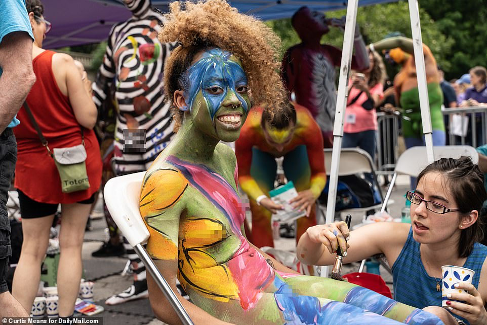 A woman smiles broadly as she is painted in a rainbow of color while sitting on a chair in New York City